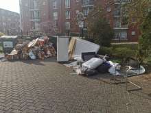 Auf dem Bild ist eine größere illegale Müllablagerung in einem Wohngebiet zu sehen. Vor mehreren schwarzen Müllcontainern stapeln sich zahlreiche Pappkartons, Verpackungen und lose Papierabfälle. Rechts daneben liegen sperrige Gegenstände wie eine alte Matratze, ein Kühlschrank, Holzbretter, schwarze Müllsäcke sowie ein umgekippter Stuhl. Im Hintergrund sind mehrstöckige Wohnhäuser mit Balkonen, grüne Büsche und ein blühender Baum zu erkennen. Die Müllmenge wirkt unkontrolliert und nimmt viel Platz ein.