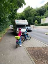 Kinder versuchen eine Verkehrsinsel zu überqueren, aber ein großes Wohnmobil versperrt die Sicht. Es fehlt die Randbegrünung und ein Baum, um Abstand für Sicht auf die Straße zu schaffen.