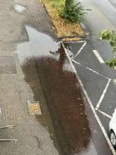 Der verstopfte Gully liegt auf dem Radweg. Auf dem Bild im Bereich, wo sich der Himmel spiegelt. 