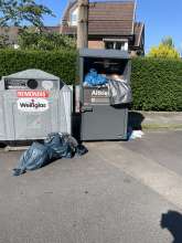 Ein Altkleidercontainer neben zwei Altglascontainern. Um den Altkleidercontainer liegen einige Tüten.